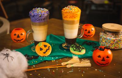 Fotografia em tons de laranja de uma mesa de madeira, duas bebidas, uma decorada com chantilly roxo e uma decorada com chantilly laranja. Em volta biscoitos decorados de abóbora, teia de aranha e elementos de halloween. Fotografia em tons de laranja de uma mesa de madeira, duas bebidas, uma decorada com chantilly roxo e uma decorada com chantilly laranja. Em volta biscoitos decorados de abóbora, teia de aranha e elementos de halloween.