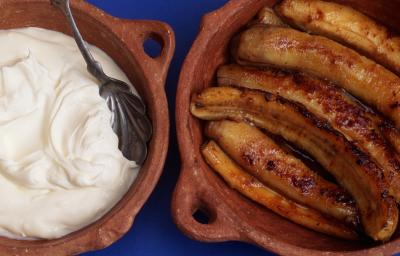 Fotografia vista de cima de bananas cortadas ao meio flambadas dentro de um recipiente marrom de cerâmica. Ao lado esquerdo, um creme branco com consistência mais densa dentro do mesmo recipiente, e dentro do creme, uma colher de prata apoiada. Fotografia vista de cima de bananas cortadas ao meio flambadas dentro de um recipiente marrom de cerâmica. Ao lado esquerdo, um creme branco com consistência mais densa dentro do mesmo recipiente, e dentro do creme, uma colher de prata apoiada.