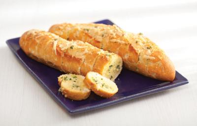 Fotografia em tons de branco, azul e amarelo de uma bancada branca com um prato quadrado azul e nele duas baguetes de pão de alho, uma delas com 2 fatias soltas.