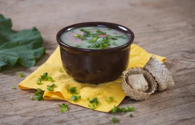 Imagem de um bowl marrom com a receita de caldo verde polvilhada com cebolinha sobre um pano amarelo e chapéus de festa junina ao lado.