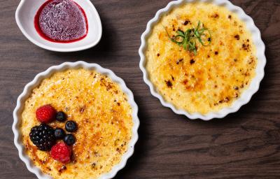 Fotografia vista de cima de dois cremes brulée, um de alho poró com queijo canastra e o outro, sabor cumaru com pistache e frutas vermelhas, os dois cremes brulée estão em ramekins. Ao lado tem uma calda de frutas vermelhas.