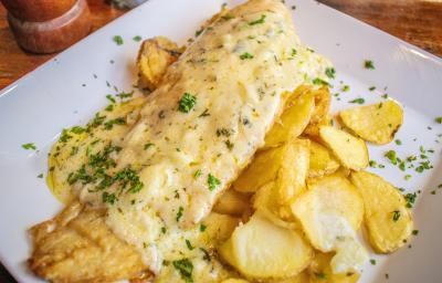 Fotografia de um filé de cação com molho de páprica, queijo e salsinha sobre batatas em rodelas finas dentro de um prato quadrado branco de vidro. O prato está sobra uma mesa de madeira. Fotografia de um filé de cação com molho de páprica, queijo e salsinha sobre batatas em rodelas finas dentro de um prato quadrado branco de vidro. O prato está sobra uma mesa de madeira.