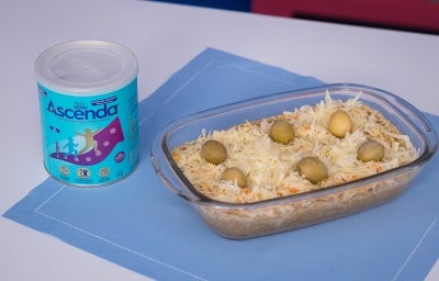 Fotografia de um recipiente refratário de vidro, retangular, ao lado da lata de Ascenda, sobre um paninho azul quadrado. Dentro do recipiente tem a torta com massa de grão de bico e ascenda, finalizada com queijo parmesão e azeitonas.