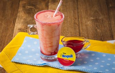 Fotografia em tons de vermelho, em uma bancada de madeira de cor mar marrom. Ao centro, um pano amarelo e um pano azul com bolinhas brancas sobrepostos. Em cima, está a bebida pink de Nesquik com um canudo dentro e ao lado, uma cápsula de Nesquik.
