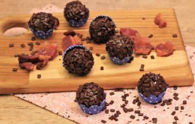 Fotografia em tons de preto em uma bancada de madeira de cor marrom. Ao centro, uma tábua de madeira contendo os brigadeiros em cima. Ao redor, há alguns pedaços de bacon espalhados. Fotografia em tons de preto em uma bancada de madeira de cor marrom. Ao centro, uma tábua de madeira contendo os brigadeiros em cima. Ao redor, há alguns pedaços de bacon espalhados.