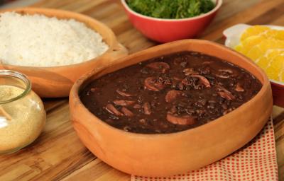 Fotografia em tons de bege em uma bancada de madeira com um pano branco com bolinhas laranjas, um recipiente de barro com a feijoada dentro. Ao lado, potes de barro com arroz, farofa e laranja dentro de cada um. Fotografia em tons de bege em uma bancada de madeira com um pano branco com bolinhas laranjas, um recipiente de barro com a feijoada dentro. Ao lado, potes de barro com arroz, farofa e laranja dentro de cada um.