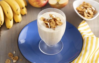 Imagem de um copo com a bebida cremosa em tom claro, decorado com cereal. Ele está sobre um prato azul e, ao redor, há cereais espalhados na mesa, uma penca de bananas, duas maçãs e um tecido listrado amarelo e branco.