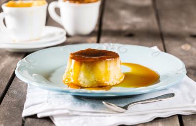 Fotografia em tons de branco e amarelo de uma bancada cinza, com um pano branco dobrado, uma colher de inox de sobremesa, um prato de sobremesa azul e nele um mini pudim redondo com calda de caramelo. Ao fundo, duas xícaras brancas com pudim dentro. Fotografia em tons de branco e amarelo de uma bancada cinza, com um pano branco dobrado, uma colher de inox de sobremesa, um prato de sobremesa azul e nele um mini pudim redondo com calda de caramelo. Ao fundo, duas xícaras brancas com pudim dentro.
