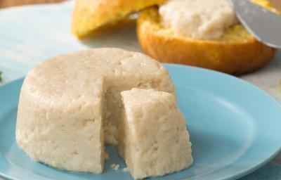 Fotografia em tons de azul e branco de uma bancada azul claro vista de frente, um prato redondo e azul contém um queijo redondo com uma fatia retirada. Ao fundo um pão redondo cortado ao meio e uma espátula com creme colocando em uma das fatias do pão. Fotografia em tons de azul e branco de uma bancada azul claro vista de frente, um prato redondo e azul contém um queijo redondo com uma fatia retirada. Ao fundo um pão redondo cortado ao meio e uma espátula com creme colocando em uma das fatias do pão.