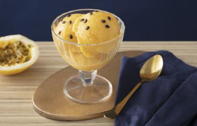 Foto em tons de amarelo da receita de sorbet de damasco e maracujá servida em uma taça de vidro sobre uma mesa de madeira com uma colher prateada ao lado sobre um pano de veludo azul. Ao fundo ainda há metade de um maracujá