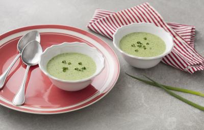 Fotografia em tons de vermelho em uma bancada de madeira cinza com um pano com listras vermelhas, um potinho branco pequeno com a sopa de alface. Ao lado, um prato vermelho grande com um potinho branco em cima dele e a sopa de alface dentro.
