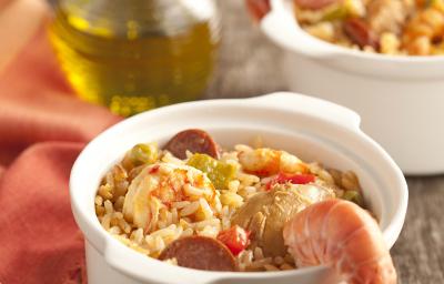 fotografia em tons de cinza e branco de uma bancada vista de frente, contém dois potinhos brancos e redondos com arroz, pedaços de carne suína e frutos do mar. Ao lado um pano laranja com dois garfos para servir e uma jarra de azeite fotografia em tons de cinza e branco de uma bancada vista de frente, contém dois potinhos brancos e redondos com arroz, pedaços de carne suína e frutos do mar. Ao lado um pano laranja com dois garfos para servir e uma jarra de azeite