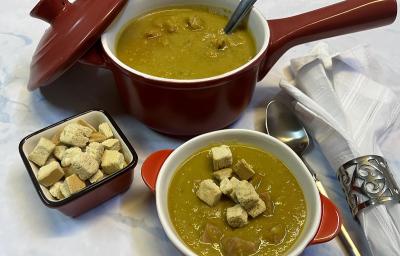 \foto em tons de verde da receita de creme de ervilha servida em uma panelinha marrom com uma colher de servir prateada dentro, à frente há um bowl laranja e branco com uma porção do creme e ao lado há crouttons e uma colher prateada \foto em tons de verde da receita de creme de ervilha servida em uma panelinha marrom com uma colher de servir prateada dentro, à frente há um bowl laranja e branco com uma porção do creme e ao lado há crouttons e uma colher prateada