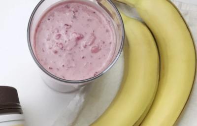 Fotografia em tons de rosa em uma mesa branca, uma toalha branca, duas bananas e um copo de vidro alto com o smoothie de morango com banana e pasta de amendoim dentro dele.