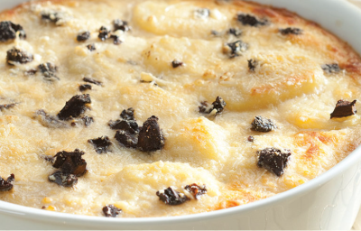 Fotografia em tons pastel, branco e dourado, ao centro refratário com batatas gratinadas com funghi sobre toalha com decoração dourada, ao fundo copos dourados e pilha de pratos brancos com guardanapo vermelho em cima. Fotografia em tons pastel, branco e dourado, ao centro refratário com batatas gratinadas com funghi sobre toalha com decoração dourada, ao fundo copos dourados e pilha de pratos brancos com guardanapo vermelho em cima.