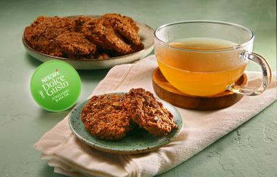 Fotografia em tons de verde em uma bancada de cimento queimado, um pano rosinha com um prato pequeno e os biscoitos de ameixa com aveia. Ao lado, uma xícara de vidro com o chá cítrico e a cápsula verde da Dolce Gusto do Chá Cítrico. Fotografia em tons de verde em uma bancada de cimento queimado, um pano rosinha com um prato pequeno e os biscoitos de ameixa com aveia. Ao lado, uma xícara de vidro com o chá cítrico e a cápsula verde da Dolce Gusto do Chá Cítrico.
