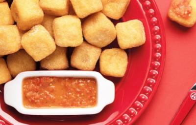 Fotografia em tons de vermelho em uma mesa vermelha, com um prato vermelho raso grande e vários dadinhos de polenta recheados com queijo coalho. Ao lado, um potinho com o molho de pimenta biquinho. Fotografia em tons de vermelho em uma mesa vermelha, com um prato vermelho raso grande e vários dadinhos de polenta recheados com queijo coalho. Ao lado, um potinho com o molho de pimenta biquinho.