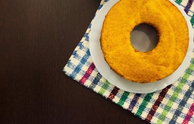 Um bolo de cenoura inteiro, redondo e simples, está colocado sobre um prato branco. A base do prato está sobre um pano de prato colorido xadrez, e o fundo é uma mesa escura de madeira, criando um contraste bonito com o bolo dourado. Um bolo de cenoura inteiro, redondo e simples, está colocado sobre um prato branco. A base do prato está sobre um pano de prato colorido xadrez, e o fundo é uma mesa escura de madeira, criando um contraste bonito com o bolo dourado.