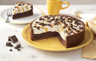 Fotografia em tons de amarelo em uma bancada branca com um prato grande amarelo e a torta de chocolate com caramelo em cima cortada ao meio. Ao lado, um prato branco com uma fatia da torta de chocolate, uma xícara amarela e pedaços de chocolates.