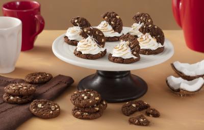 Foto de uma bancada com canecas vermelha e brancas, um tecido marrom, pedaços de coco seco. Ao centro há um prato com as receitas de cookies com merengue de coco e mais alguns Cookies Prestígio espalhados na bancada Foto de uma bancada com canecas vermelha e brancas, um tecido marrom, pedaços de coco seco. Ao centro há um prato com as receitas de cookies com merengue de coco e mais alguns Cookies Prestígio espalhados na bancada