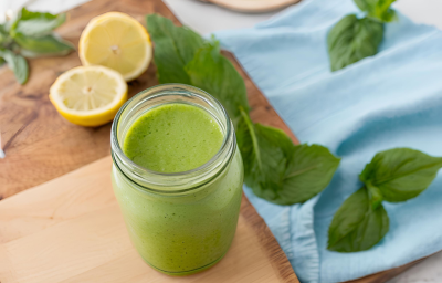 Fotografia de um copo de vidro decorativo com suco detox de cor verde feito com couve, maçã verde, pepino, gengibre e nutren. O copo está sobre uma tábua de madeira, ao lado de folhas de hortelã e metades de limão siciliano sobre a mesa. Fotografia de um copo de vidro decorativo com suco detox de cor verde feito com couve, maçã verde, pepino, gengibre e nutren. O copo está sobre uma tábua de madeira, ao lado de folhas de hortelã e metades de limão siciliano sobre a mesa.