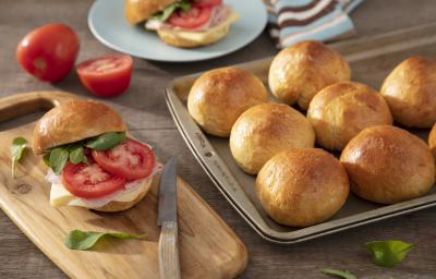 Fotografia em tons de marrom em uma bancada de madeira escura, uma tábua de madeira com um sanduíche de pão de batata doce recheado com presunto, queijo e rodela de tomate. Ao lado, uma forma com vários pãezinhos.