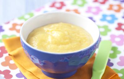 Fotografia em tons de azul em uma bancada de madeira azul clara, um pano com flores coloridas, um paninho laranja, um recipiente azul com a papinha dentro dele. Ao lado, uma colher infantil verde.