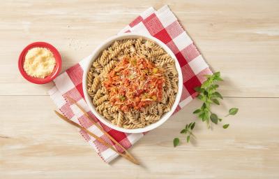 Fotografia em tons de vermelho em uma bancada de madeira com um pano de xadrez vermelho, em cima um prato fundo redondo com o macarrão integral com molho de frango dentro. Ao lado, um pegador de macarrão e um potinho vermelho com queijo ralado. Fotografia em tons de vermelho em uma bancada de madeira com um pano de xadrez vermelho, em cima um prato fundo redondo com o macarrão integral com molho de frango dentro. Ao lado, um pegador de macarrão e um potinho vermelho com queijo ralado.