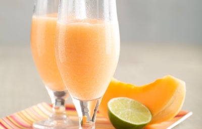 A imagem mostra duas taças com um smoothie de cor laranja claro, possivelmente feito de melão. As taças estão sobre uma bandeja colorida com listras, que adiciona um toque vibrante. Ao lado, há uma fatia de melão e um limão cortado ao meio