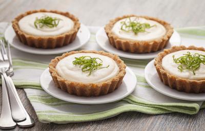 Fotografia em tons de verde em uma mesa de madeira com um pano listrado em verde e quatro pratinhos brancos cada um com a torta creme de limão. Ao lado, três garfos.