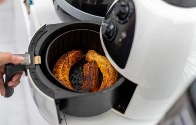 Fotografia dem tons claros de uma mão puxando a cestinha da air fryer branca com uma receita de banana douradinha. Fotografia dem tons claros de uma mão puxando a cestinha da air fryer branca com uma receita de banana douradinha.