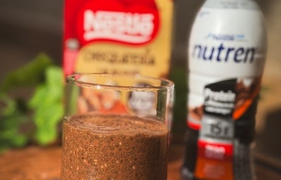 Fotografia de um copo de vidro com chia pudding feita com NUTREN Protein sabor chocolate. O copo está sobre uma tábua grande de madeira, ao fundo tem uma garrafinha de NUTREN e uma embalagem de NESTLÉ Chocolateria.