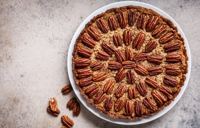 Fotografia vista de cima de uma torta inteira de nozes e doce de leite, apoiada em um prato grande, raso e branco sobre uma mesa tipo mármore. Ao lado do prato tem seis nozes sobre a mesa.