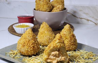 Foto da receita de coxinha de estrogonofe empanada em batata palha, servidas em um prato cinza, com potes ao fundo contendo ketchup e mostarda. Foto da receita de coxinha de estrogonofe empanada em batata palha, servidas em um prato cinza, com potes ao fundo contendo ketchup e mostarda.