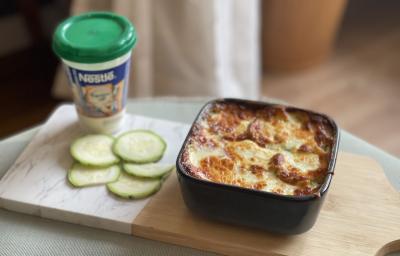 Fotografia em tons de marrom e verde em uma bancada de madeira, com uma tábua de madeira e em cima um pote quadrado com a lasanha de abobrinha com frango e requeijão light. Ao lado, um pote de requeijão nestlé. Fotografia em tons de marrom e verde em uma bancada de madeira, com uma tábua de madeira e em cima um pote quadrado com a lasanha de abobrinha com frango e requeijão light. Ao lado, um pote de requeijão nestlé.