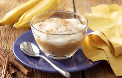 Fotografia em tons de branco em uma bancada de madeira de cor marrom. Ao centro, um prato azul contendo um recipiente com a banoffee. Ao lado, há uma colher, um pano amarelo e ao fundo, 2 bananas. Fotografia em tons de branco em uma bancada de madeira de cor marrom. Ao centro, um prato azul contendo um recipiente com a banoffee. Ao lado, há uma colher, um pano amarelo e ao fundo, 2 bananas.
