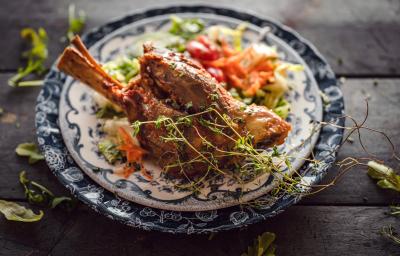 Fotografia de um cordeiro temperado e assado com vinho e ervas e um prato azul e branco, e embaixo da carne, uma salada colorida. Ao redor do prato, algumas folhas de ervas sobre uma mesa de madeira em tom escuro.