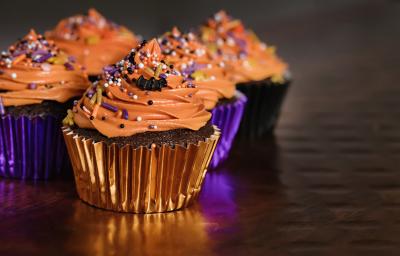 Foto de cupcakes cobertos por um creme laranja, decorados com confeitos coloridos em embalagens brilhantes nos tons de roxo e laranja.