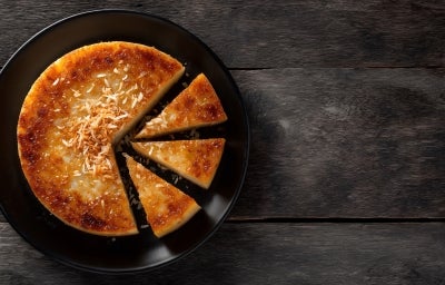 Um bolo de mandioca caramelizado em formato redondo está servido em um prato preto. Algumas fatias já foram cortadas, revelando sua textura cremosa. A cobertura tem um tom dourado e está decorada com coco tostado no centro.
