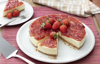 Fotografia em tons de vermelho, em uma bancada de madeira de cor marrom. No centro, uma toalha vermelha e em cima um prato redondo branco contendo o cheesecake. Ao fundo, um pires branco, contendo uma fatia do cheesecake com 2 morangos e um garfo ao lado. Fotografia em tons de vermelho, em uma bancada de madeira de cor marrom. No centro, uma toalha vermelha e em cima um prato redondo branco contendo o cheesecake. Ao fundo, um pires branco, contendo uma fatia do cheesecake com 2 morangos e um garfo ao lado.