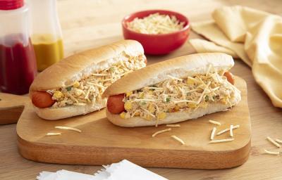 Foto da receita de cachorro quente cremoso. Observa-se dois cachorros quentes em cima de uma tábua com o recheio de frango por cima da salsicha e, para finalizar, batata palha. Ao fundo, dois potinhos de ketchup e mostarda decoram a foto Foto da receita de cachorro quente cremoso. Observa-se dois cachorros quentes em cima de uma tábua com o recheio de frango por cima da salsicha e, para finalizar, batata palha. Ao fundo, dois potinhos de ketchup e mostarda decoram a foto