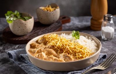 Fotografia em tons de cinza com um prato fundo ao centro. Dentro do prato existe uma receita de frango com cubos com molho de cor rosé, batata palha e arroz. Ao fundo existe um potinho com sal. Fotografia em tons de cinza com um prato fundo ao centro. Dentro do prato existe uma receita de frango com cubos com molho de cor rosé, batata palha e arroz. Ao fundo existe um potinho com sal.