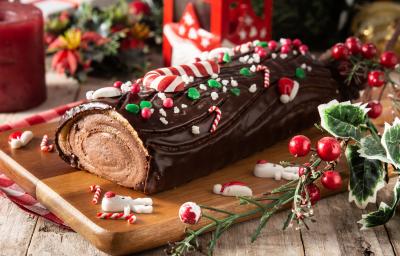 fotografia em tons de marrom, verde e vermelho tirada de uma tábua de madeira. Por cima contém um rocambole de chocolate em formate de troco de arvore com enfeites de Natal.