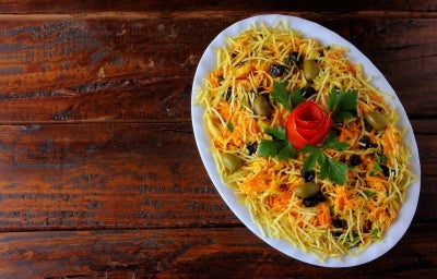 Fotografia de uma salada apresentada em um prato oval branco, com ingredientes finamente picados em tons amarelos e laranjas, azeitonas verdes, batata palha e um enfeite de tomate em forma de rosa, rodeado por folhas de salsa fresca. Fotografia de uma salada apresentada em um prato oval branco, com ingredientes finamente picados em tons amarelos e laranjas, azeitonas verdes, batata palha e um enfeite de tomate em forma de rosa, rodeado por folhas de salsa fresca.