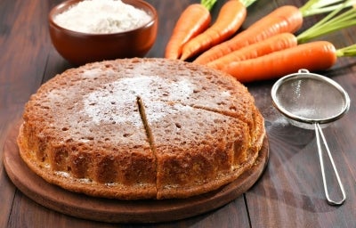 Imagem apresenta um bolo de cenoura em uma tábua de madeira, com uma crosta dourada e uma fatia cortada, revelando seu interior úmido. Cenouras frescas e uma tigela de farinha estão ao fundo, destacando a natureza caseira e apetitosa do bolo. Imagem apresenta um bolo de cenoura em uma tábua de madeira, com uma crosta dourada e uma fatia cortada, revelando seu interior úmido. Cenouras frescas e uma tigela de farinha estão ao fundo, destacando a natureza caseira e apetitosa do bolo.