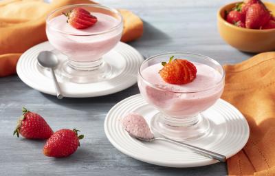 Imagem aproximada da receita de Mousse de Morango Zero Lactose, em tom rosa claro, servida em potes de vidro, decoradas com morangos fatiados. A receita está sobre pratos brancos em uma bancada com um tecido laranja e mais morangos inteiros decorando Imagem aproximada da receita de Mousse de Morango Zero Lactose, em tom rosa claro, servida em potes de vidro, decoradas com morangos fatiados. A receita está sobre pratos brancos em uma bancada com um tecido laranja e mais morangos inteiros decorando