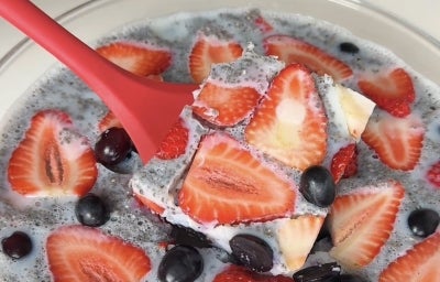 Fotografia em tons claros de uma espátula vermelha segurando uma porção da gelatina de frutas e chia, que está dentro de um recipiente de vidro.