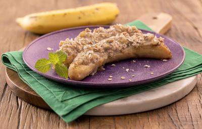 Foto da receita de banana grelhada recheada servida em um prato roxo sobre um paninho verde em uma tábua de madeira de forma circular. Ao fundo há uma banana para decoração Foto da receita de banana grelhada recheada servida em um prato roxo sobre um paninho verde em uma tábua de madeira de forma circular. Ao fundo há uma banana para decoração