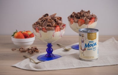 Foto da receita de pavê de leite moça com morangos e chocolate, servidos em taças com uma base azul, sobre uma bancada de madeira decorada com um tecido, pedaços de chocolate, um pote com morangos e uma lata de Leite Moça Foto da receita de pavê de leite moça com morangos e chocolate, servidos em taças com uma base azul, sobre uma bancada de madeira decorada com um tecido, pedaços de chocolate, um pote com morangos e uma lata de Leite Moça