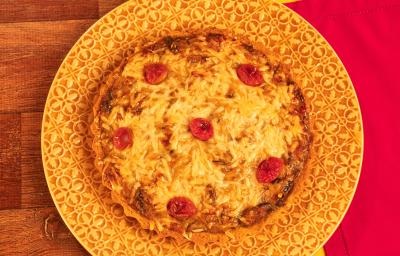 Fotografia mostra uma quiche com queijo gratinado e pedaços de tomate sobre um prato amarelo. Ao fundo, uma mesa de madeira e um pano vermelho. Fotografia mostra uma quiche com queijo gratinado e pedaços de tomate sobre um prato amarelo. Ao fundo, uma mesa de madeira e um pano vermelho.